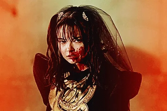 GIF of lachesis from kamen rider gotchard in her TTFC special. she's slowly shifting to the side and looking off camera, her face covered in blood. the image is highly saturated in warm tones, with hot red fading in on each side of her